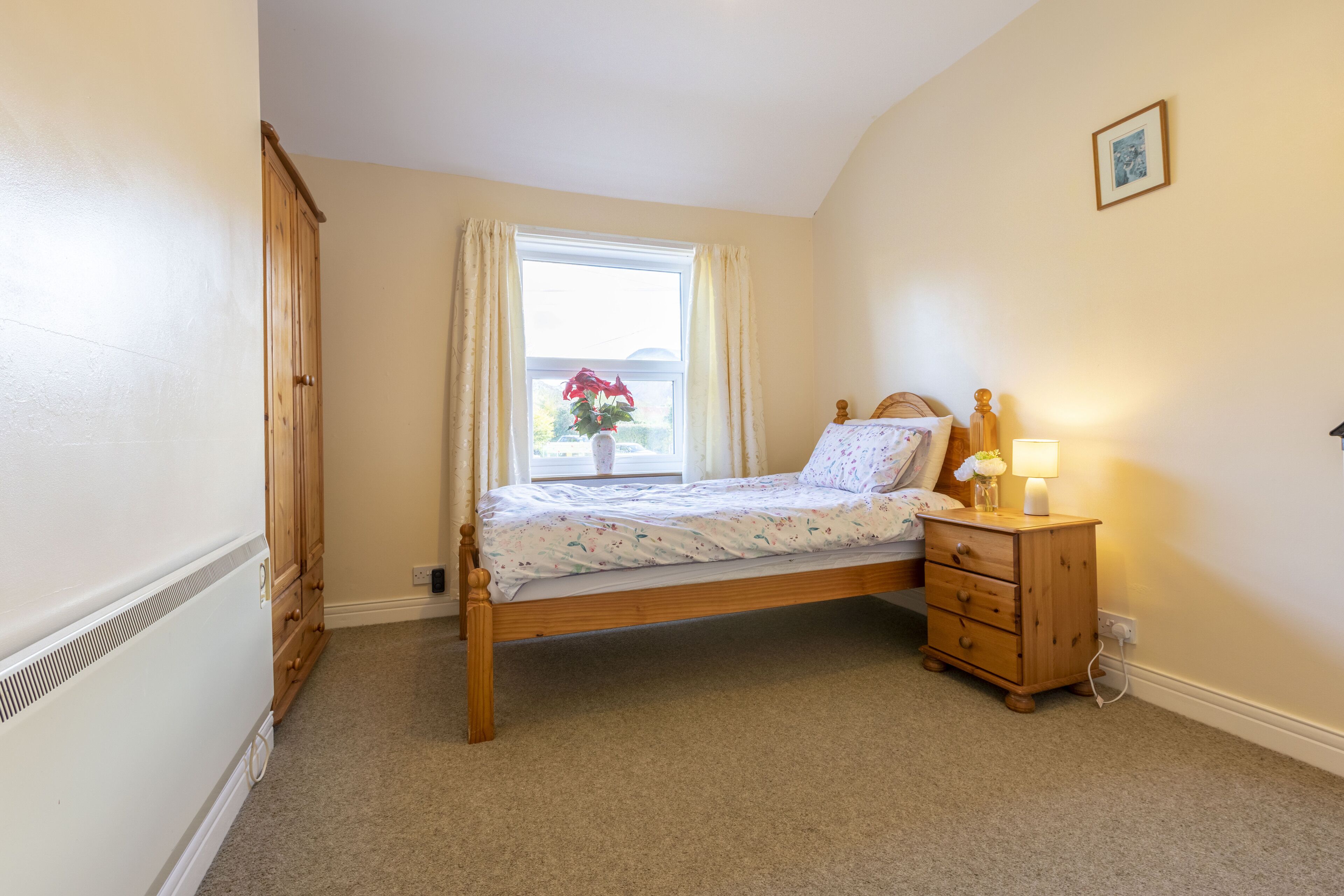 A bedroom with a wooden bed, a bedside table with a lamp, a window with curtains, and a radiator.