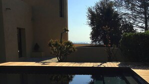 A wooden deck with a red table and chairs, a swimming pool, and a building with a view of the landscape. - New: Villa 200 m2 with sea view and heated swimming pool, 4 bedrooms. (Porto-Vecchio)