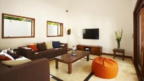 A living room with a sofa, coffee table, and orange bean bag. There is a flat-screen TV mounted on the wall and a small table with a lamp.