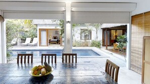 A dining area with a table and chairs, a bowl of fruit, and a view of a swimming pool and outdoor seating.