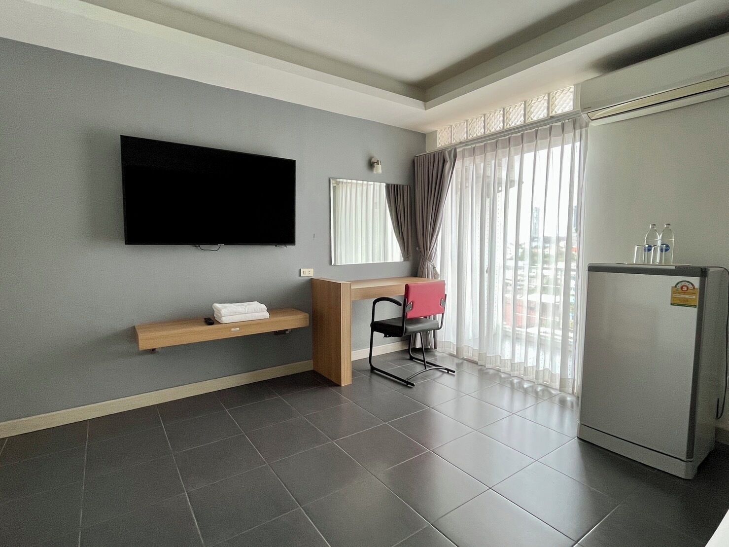 Habitación de hotel con televisor de pantalla plana montado en la pared, un escritorio de madera con una silla, un estante con una toalla, un refrigerador y un ventanal con cortinas traslúcidas.