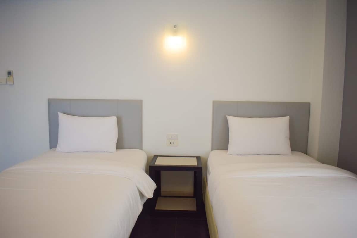 A hotel room with two single beds, each with a headboard and a pillow. There is a small bedside table between the beds.