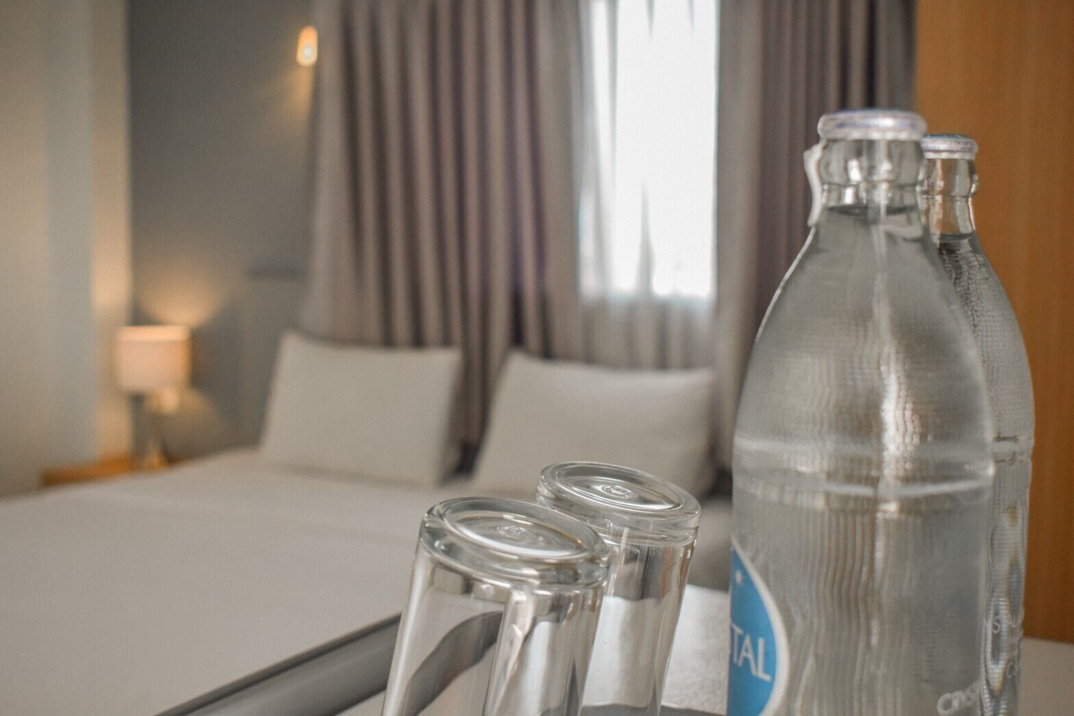 Una habitación de hotel con una cama, una mesita de noche, una lámpara y una mesa con una botella y vasos.