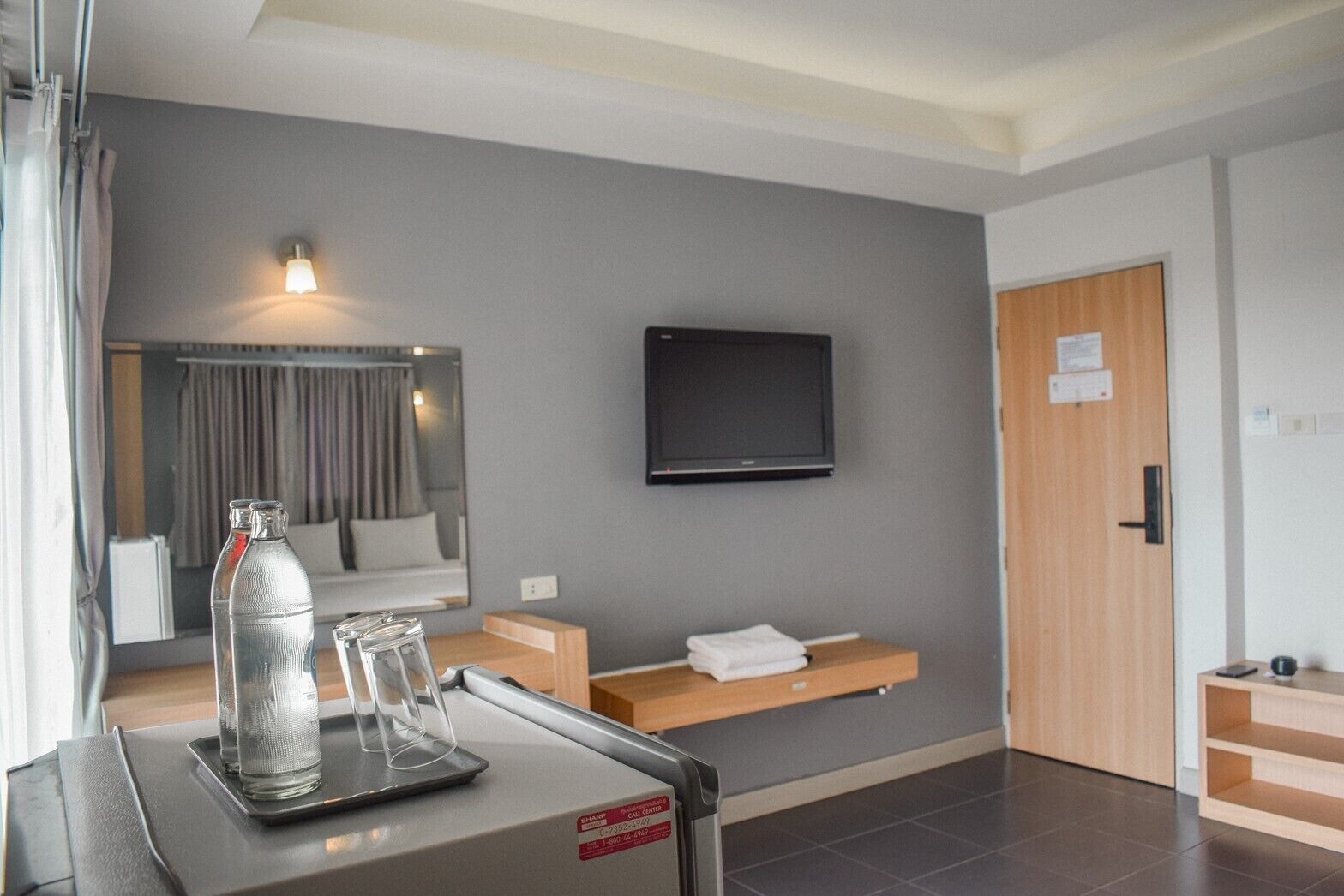 A modern hotel room with a bed, a TV mounted on the wall, a small table with a bottle and glasses, and a wooden door.
