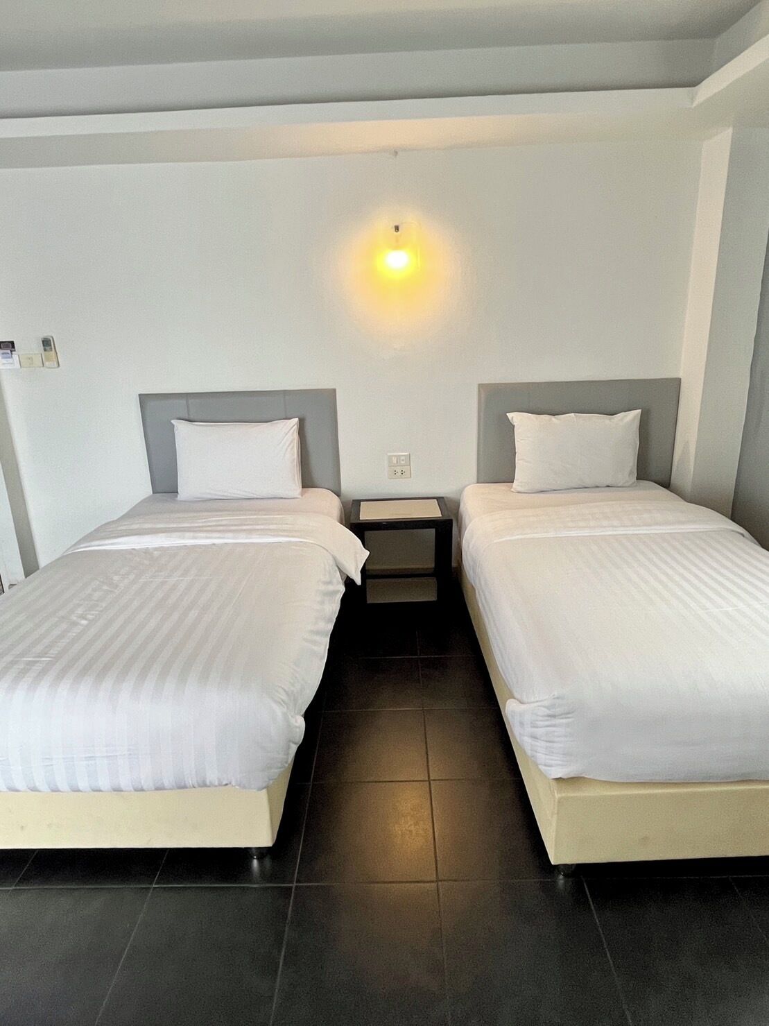 Una habitación de hotel con dos camas, una mesita de noche, una lámpara fijada a la pared y suelo de baldosas.