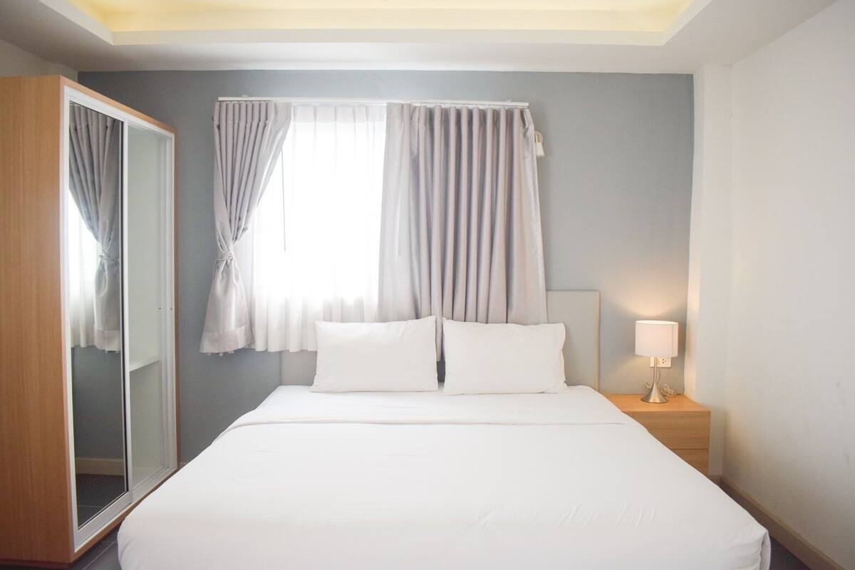 A hotel room with a bed, a nightstand with a lamp, a window with curtains, and a wardrobe.