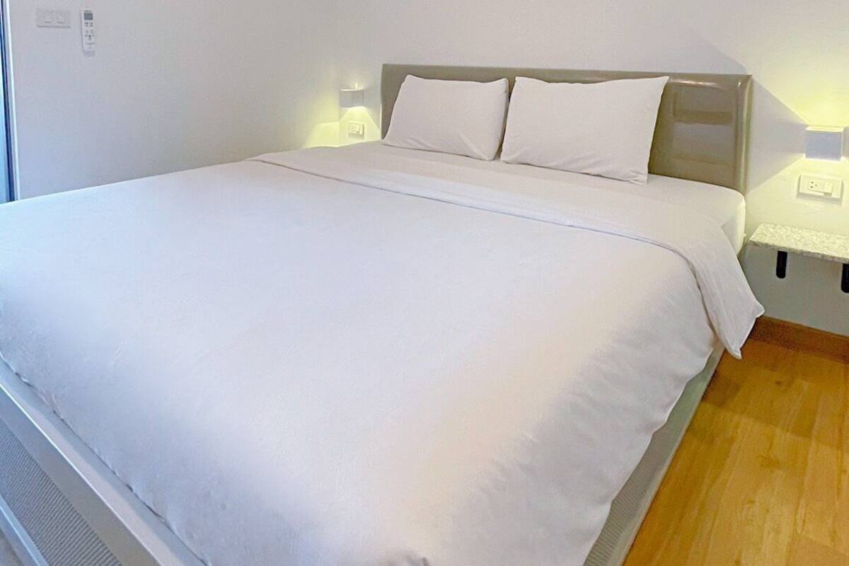 Una cama bien hecha en una habitación de paredes blancas y suelo de madera. Hay dos mesitas de noche con lámparas, y se ve un enchufe en la pared.