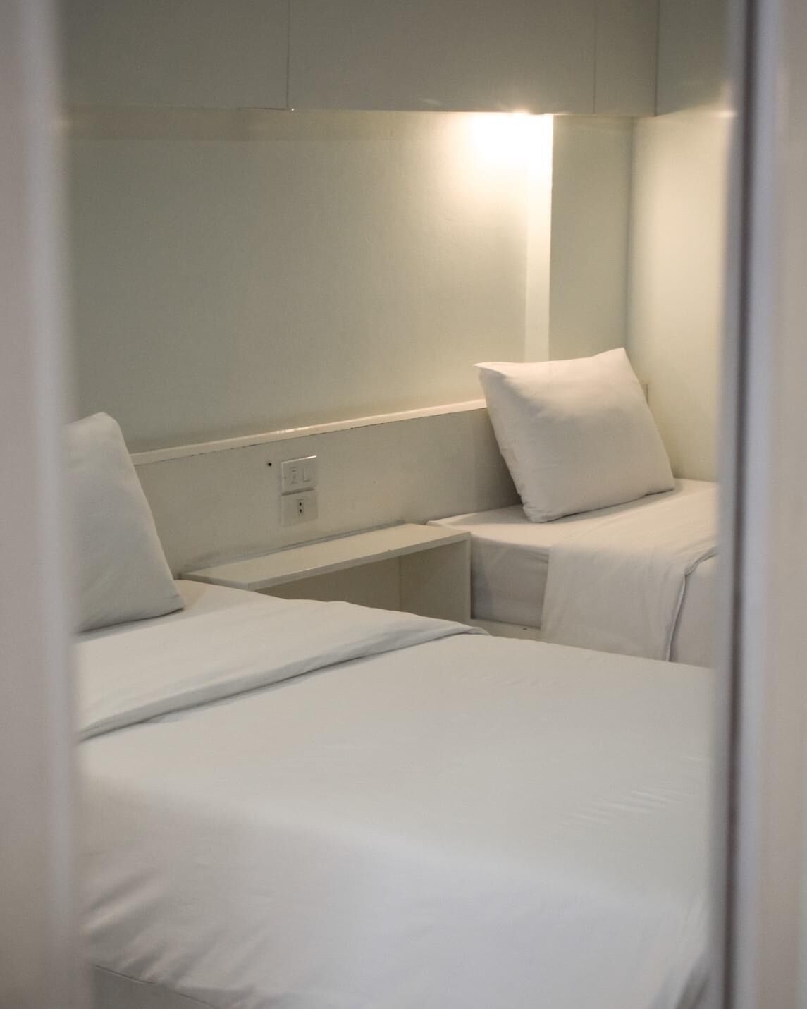 A small, compact room with two single beds, a bedside table, and a wall-mounted light.