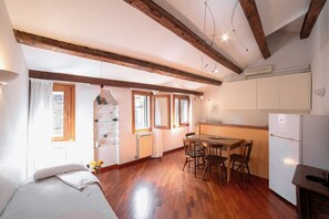 A compact room with a bed, a dining table with chairs, a small kitchen area with a refrigerator, and wooden beams on the ceiling.