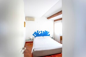 Une chambre à coucher avec un lit, un plancher en bois, des murs blancs, un ventilateur de plafond et une fenêtre avec des rideaux.