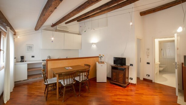 A spacious living area with a wooden dining table and chairs, a small white refrigerator, a television, and a kitchen area with cabinets and a sink. - Ca' Marina, a few steps from the Biennale di Venezia (Venezia)