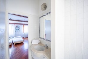A hotel room with a wooden floor, a bed, a bathroom with a sink and mirror, and a view of another room with a bed and a window.