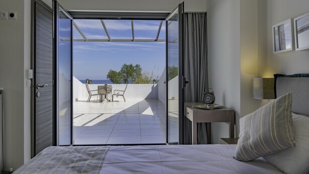 A bedroom with a large bed, a nightstand, a lamp, and a view of an outdoor patio with a table and chairs. - Blue oyster Beachfront villa, No car needed 1 (Rethymno)