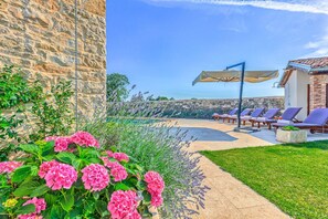 Un mur de pierre, un jardin parsemé de fleurs roses et un espace piscine avec des chaises longues et des parasols.