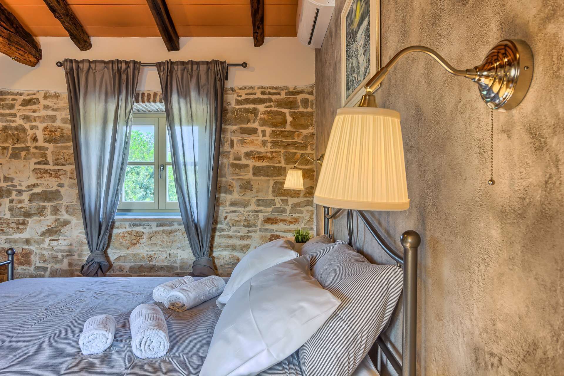 A bedroom with a stone wall, a bed with rolled towels, a lamp, and a window with curtains.
