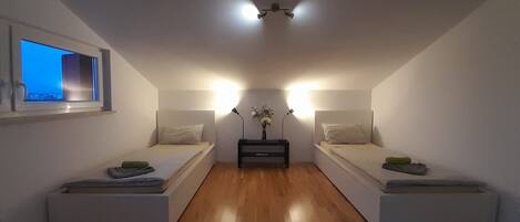 A small, modern room with two single beds, a nightstand with a lamp, and a window.