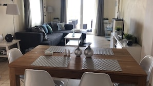 Una sala de estar moderna con una mesa de comedor de madera, sillas blancas, un sofá oscuro y un ventanal con cortinas.