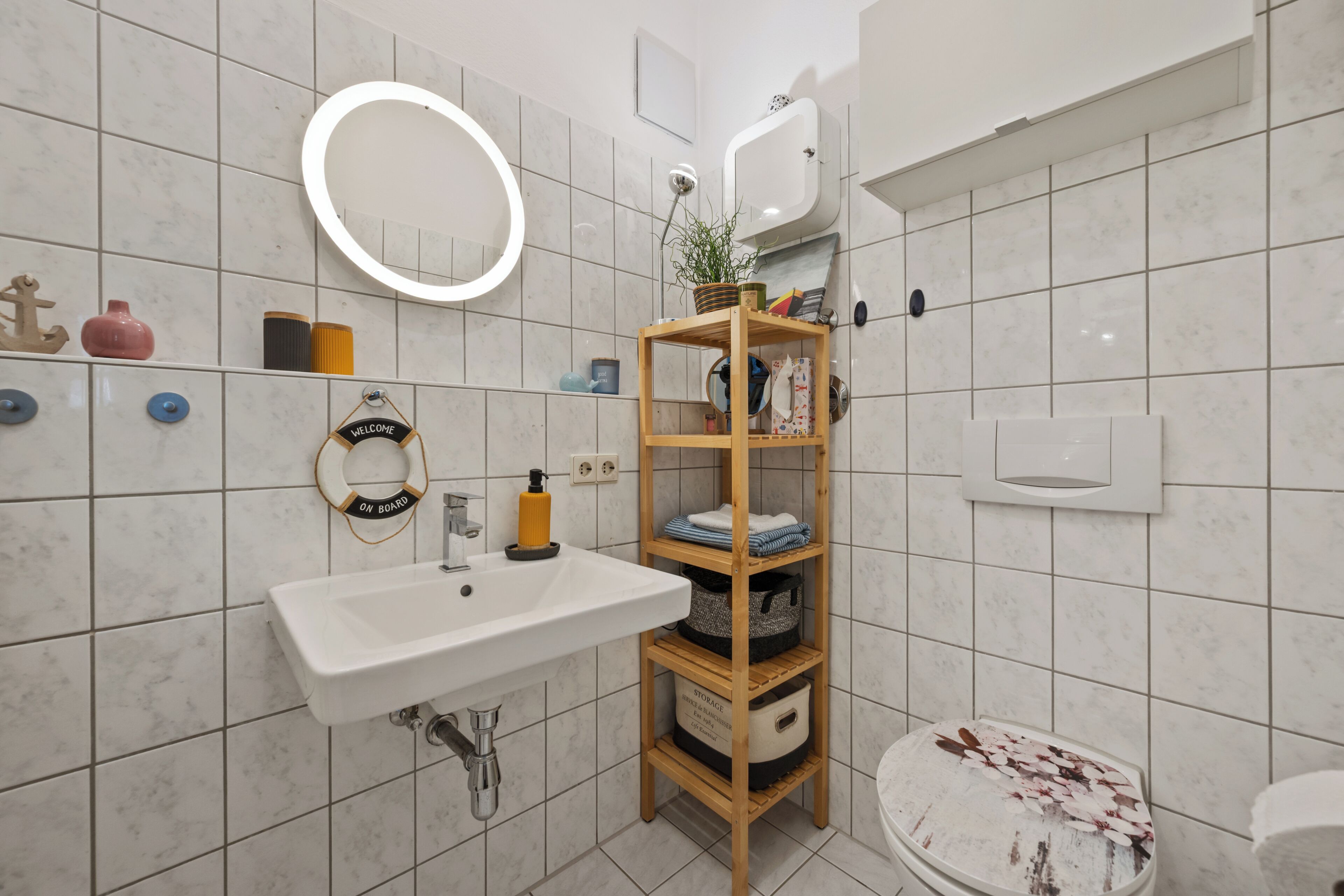 A bathroom with a white sink, a wooden shelf with storage, a toilet, and a round mirror.