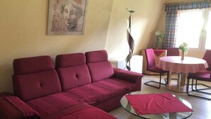 Un salon avec un canapé rouge, une table basse en verre, un tableau au mur et un coin repas avec une nappe à carreaux.