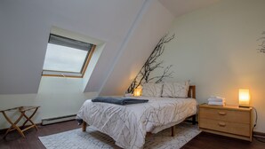 Une chambre avec un plafond en pente, un lit recouvert d’une couvre-lit à motifs, une table de chevet avec une lampe et une fenêtre donnant sur le ciel.
