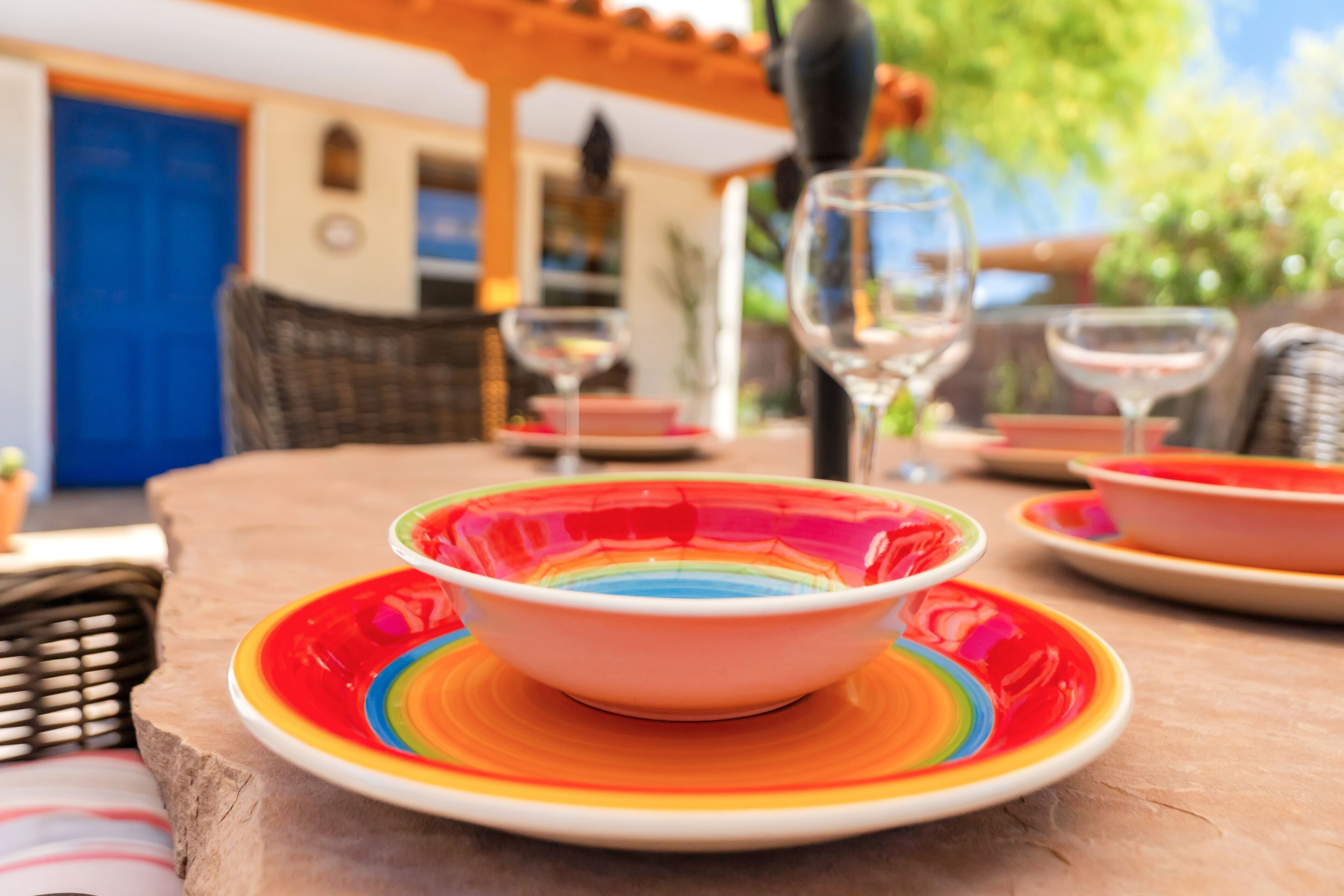 A table with colorful plates and a bowl, a wicker chair, and a blue door in the background.
