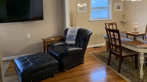 A living room with a black leather sofa and ottoman, a wooden dining table with chairs, a chandelier, and a TV mounted on the wall. - Quiet, Charming Lake Front Home with Kayaks, 2 Guest Maximum (Hot Springs)