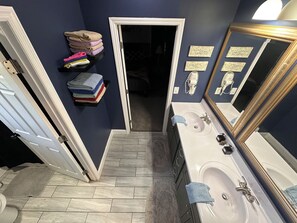 A bathroom with two sinks, a mirror, towels on a rack, and a door leading to another room.