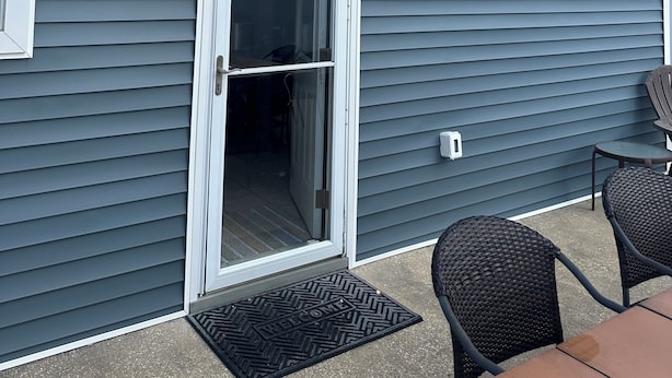Une maison avec un revêtement extérieur gris, une porte-fenêtre coulissante et un petit espace patio comprenant deux chaises en osier et une table.