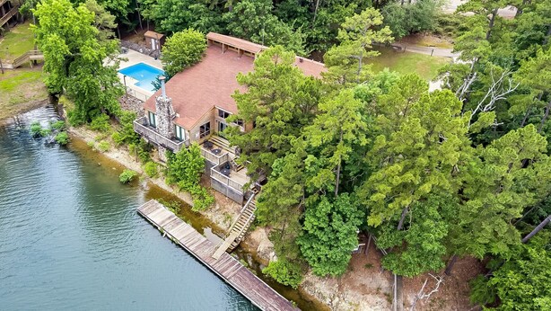 An aerial view of a lakeside property with a swimming pool, wooden deck, and a stone house.