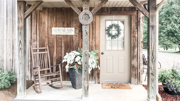 A rustic barn-style building with a wooden exterior, a sign reading "Stony Hill Barn Bed and Breakfast," a welcome mat, and a rocking chair outside.