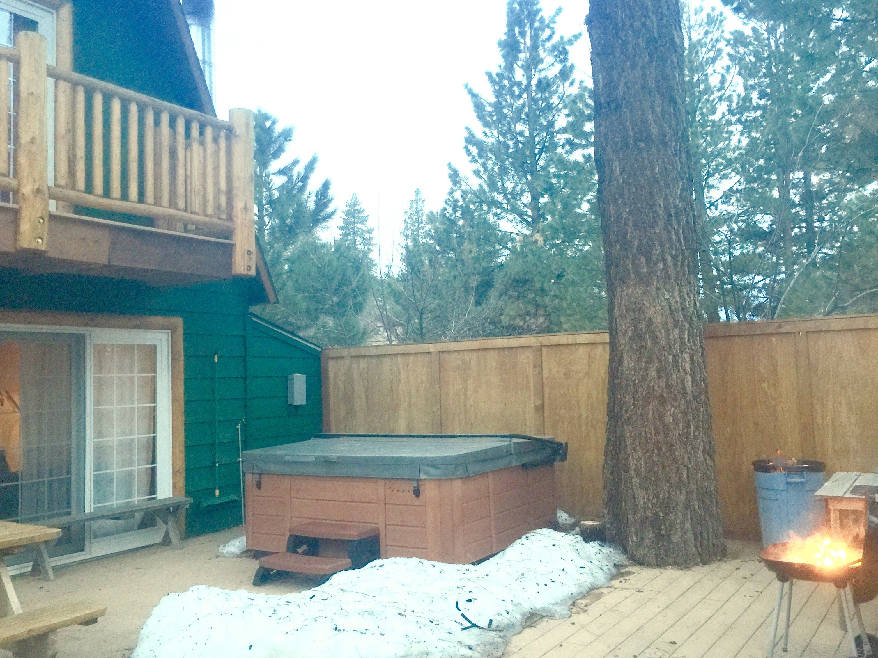 A backyard with a hot tub, a wooden deck, a wooden fence, and a tree.