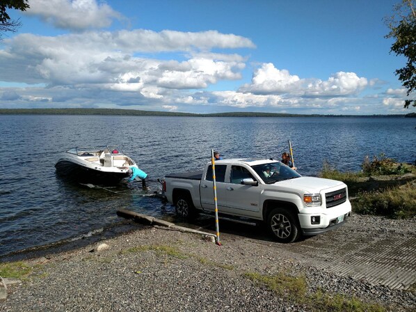 Una camioneta blanca estacionada en un muelle de grava junto a un lago, con un bote amarrado a ella.