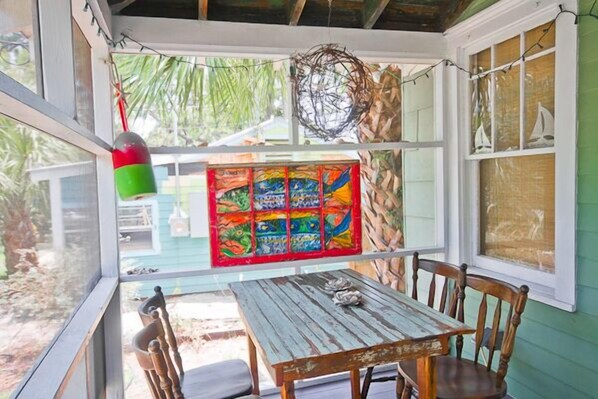A covered patio with a wooden table and chairs, a colorful artwork on the wall, and a hanging nest.