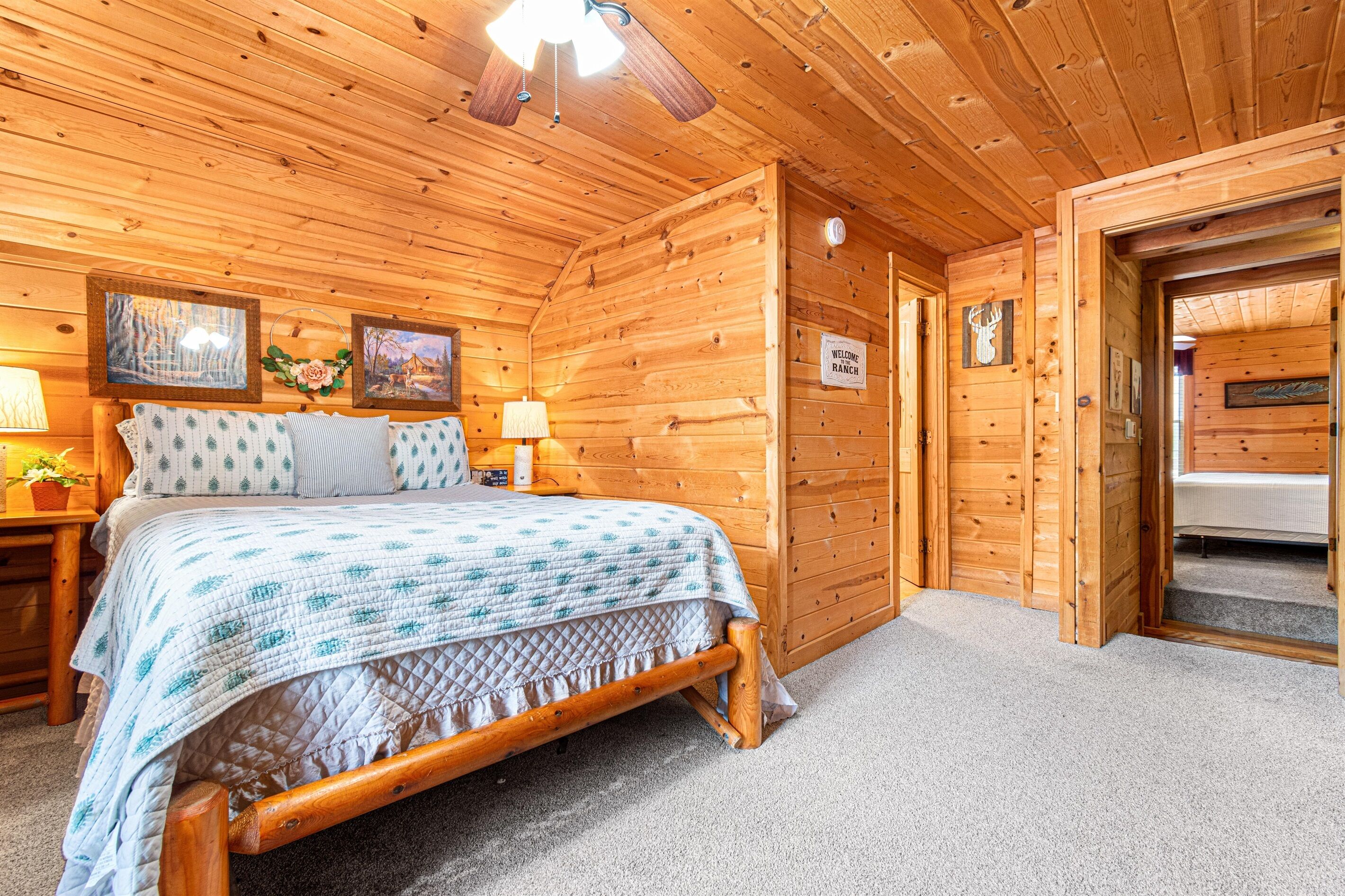A wooden cabin bedroom with a bed, a nightstand, a lamp, framed pictures, and a 'Welcome Ranch' sign.