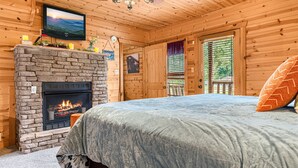 A cozy log cabin room with a stone fireplace, a bed with a blue bedspread, a television mounted on the wall, and a chandelier. - End of Summer Retreat Stunning Views, Horses, Pool (Sevierville)