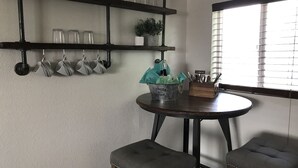 A kitchen with a round table, two stools, a shelf with glassware, and a window with blinds. - Cut Bank Unit #1 near Glacier National Park! (Cut Bank)