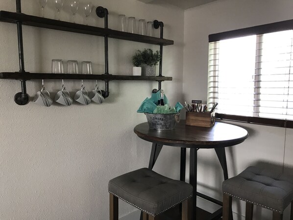 Una cocina con una mesa redonda, dos taburetes, una repisa con cristalería y una ventana con persianas.
