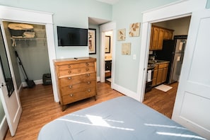 A bedroom with a bed, a wooden dresser, a closet with a TV, and a kitchen area visible through an open door. - Inn The Heights - Apt 2 (Tampa)