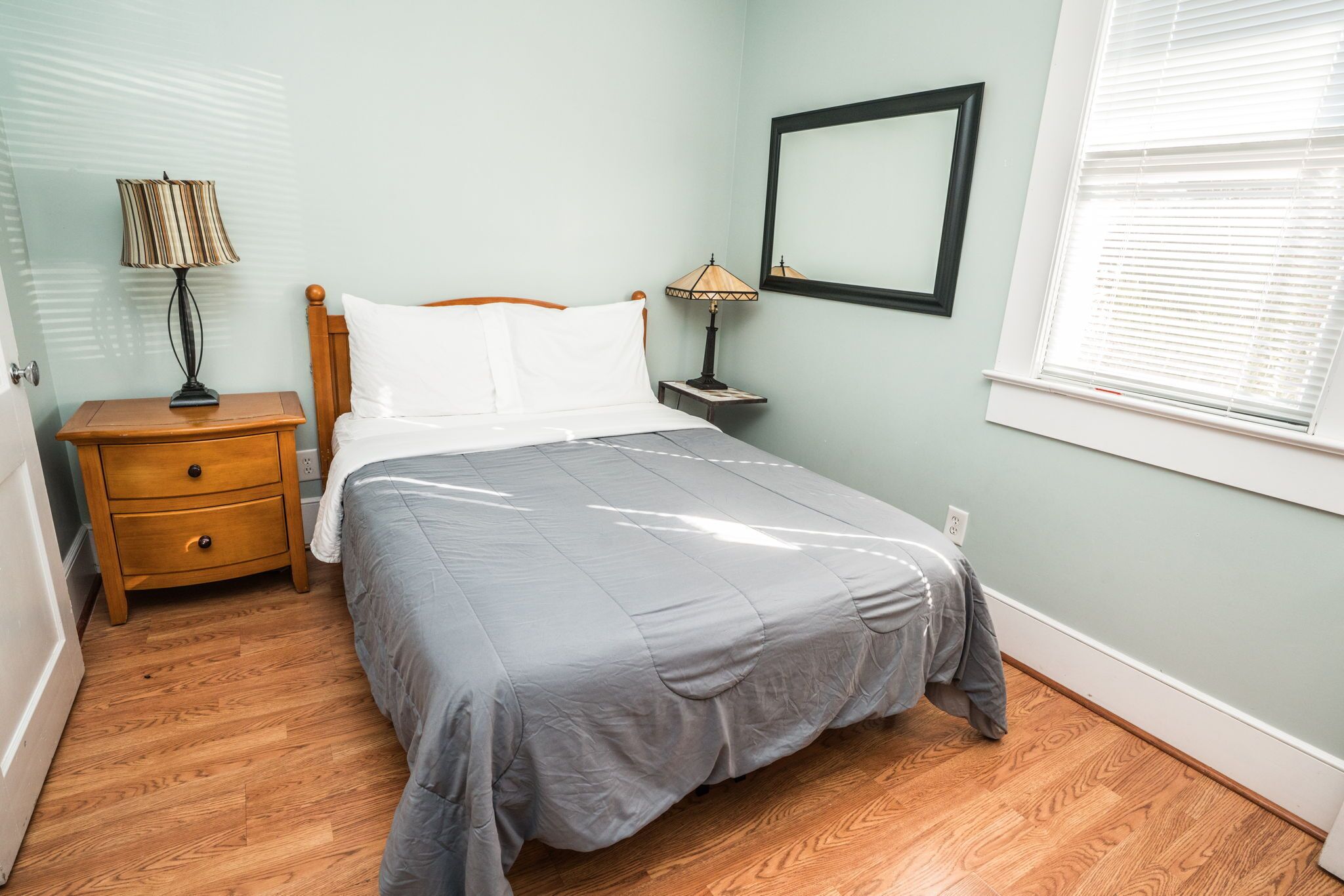A neatly arranged bedroom with a bed, two bedside tables, a lamp, a mirror, and a window with blinds.