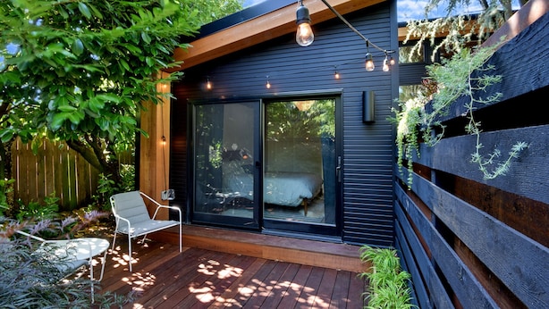 Une terrasse extérieure moderne avec un plancher en bois, une chaise en métal blanche et une porte vitrée donnant sur une pièce où se trouve un lit.