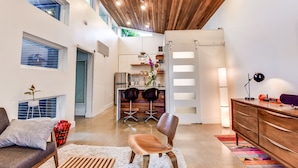 Un intérieur moderne avec un plafond haut, des poutres en bois, un coin cuisine et un espace salon comprenant un canapé et des fauteuils.