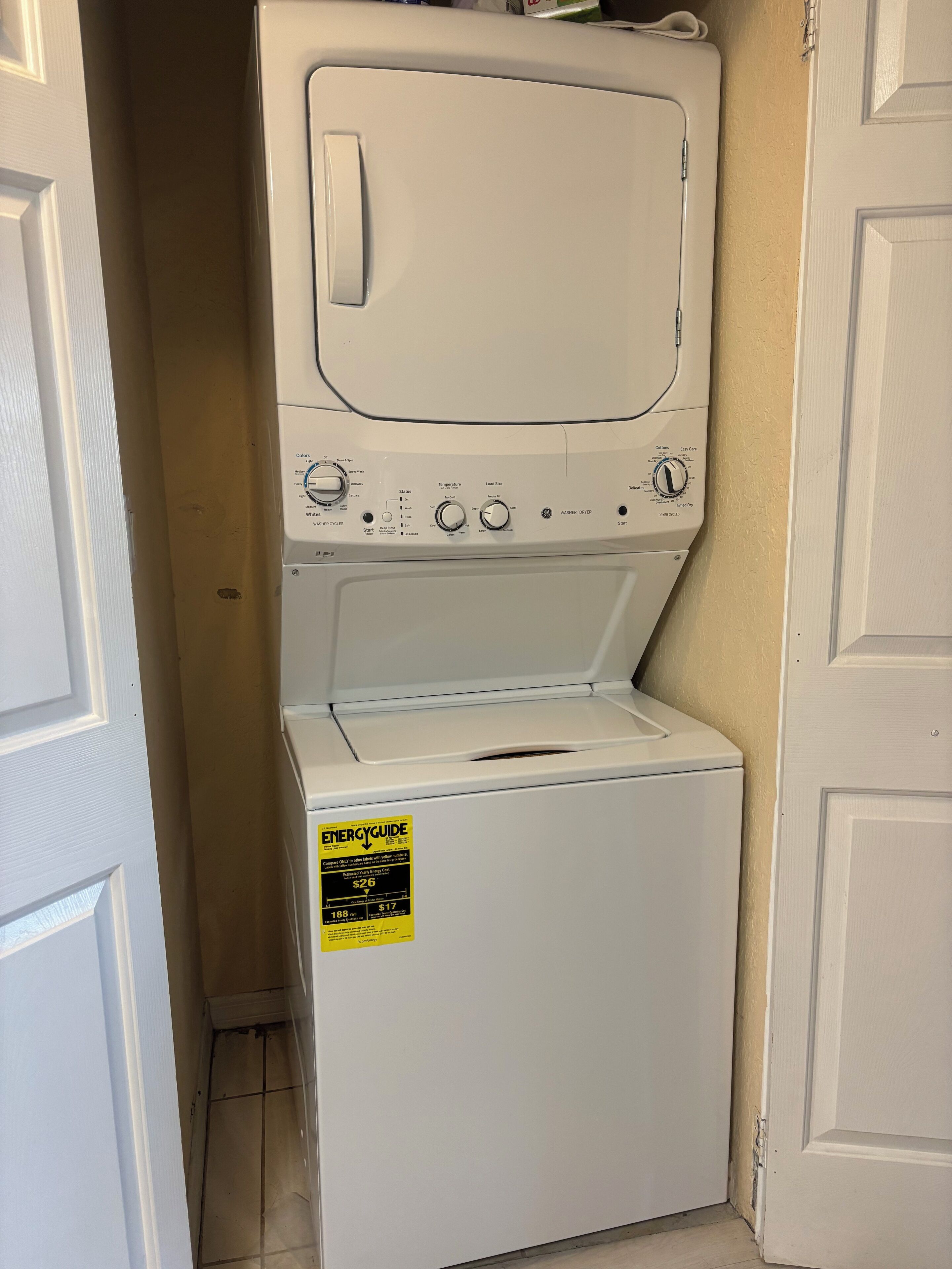 Un appareil combiné lave-linge et sèche-linge à chargement frontal, de couleur blanche, muni d’une étiquette indiquant sa consommation énergétique, situé dans un couloir étroit avec des portes blanches.
