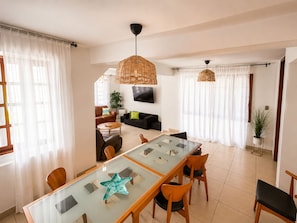 A modern living room with a glass dining table, a sofa, and a TV mounted on the wall.