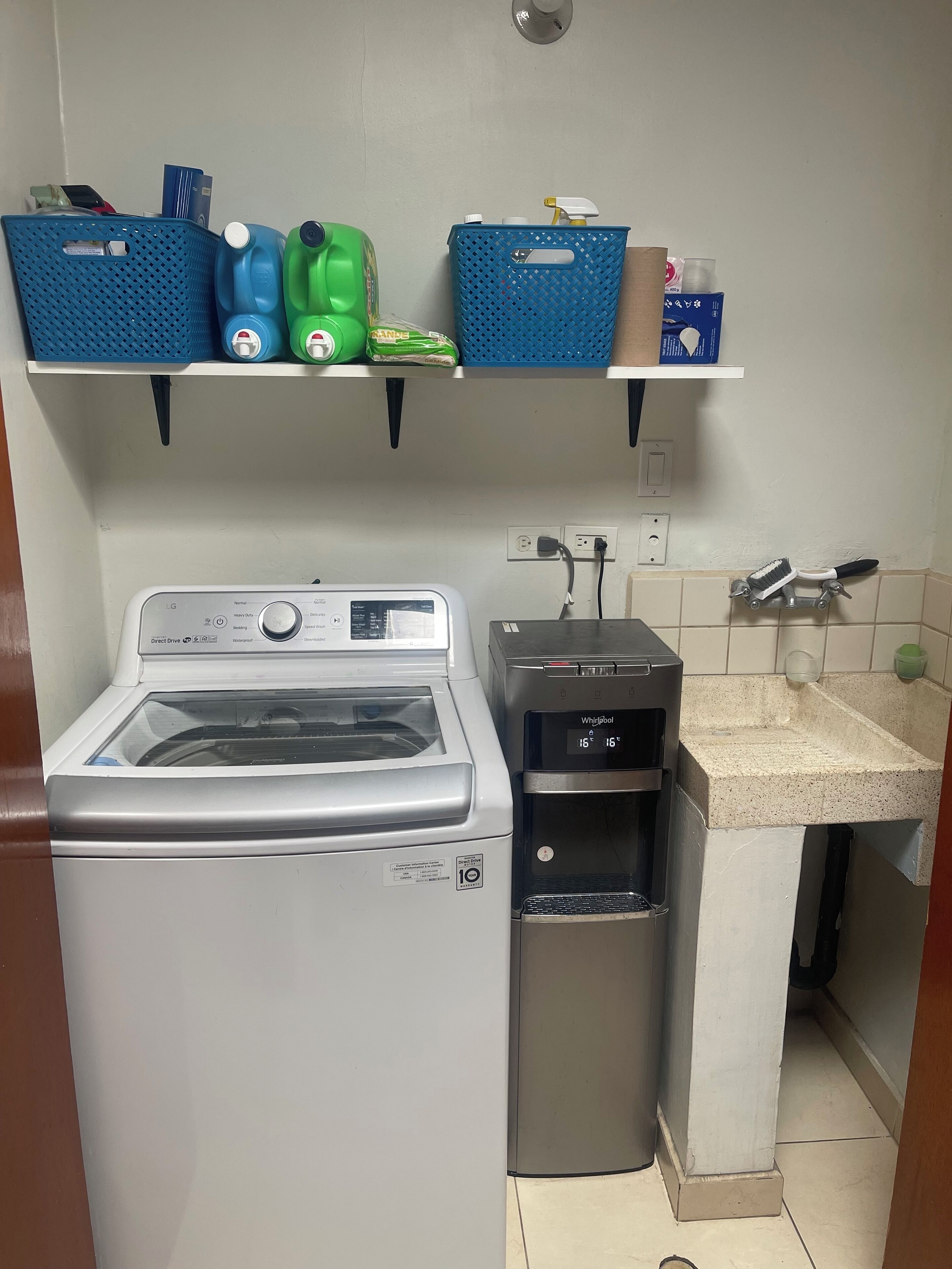A laundry area with a front-loading washer and dryer, a water dispenser, and shelves with cleaning supplies.