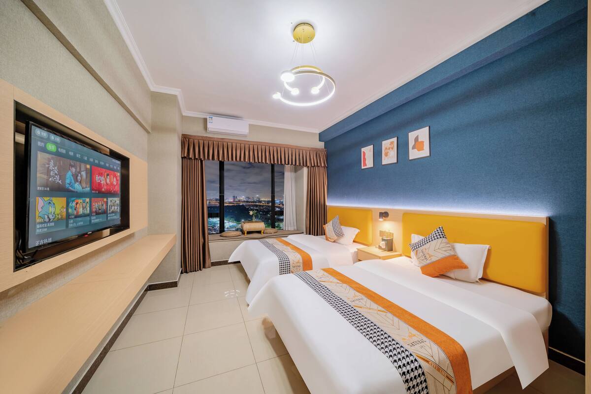 A hotel room with two beds, a TV on the wall, a city view, and a blue accent wall.