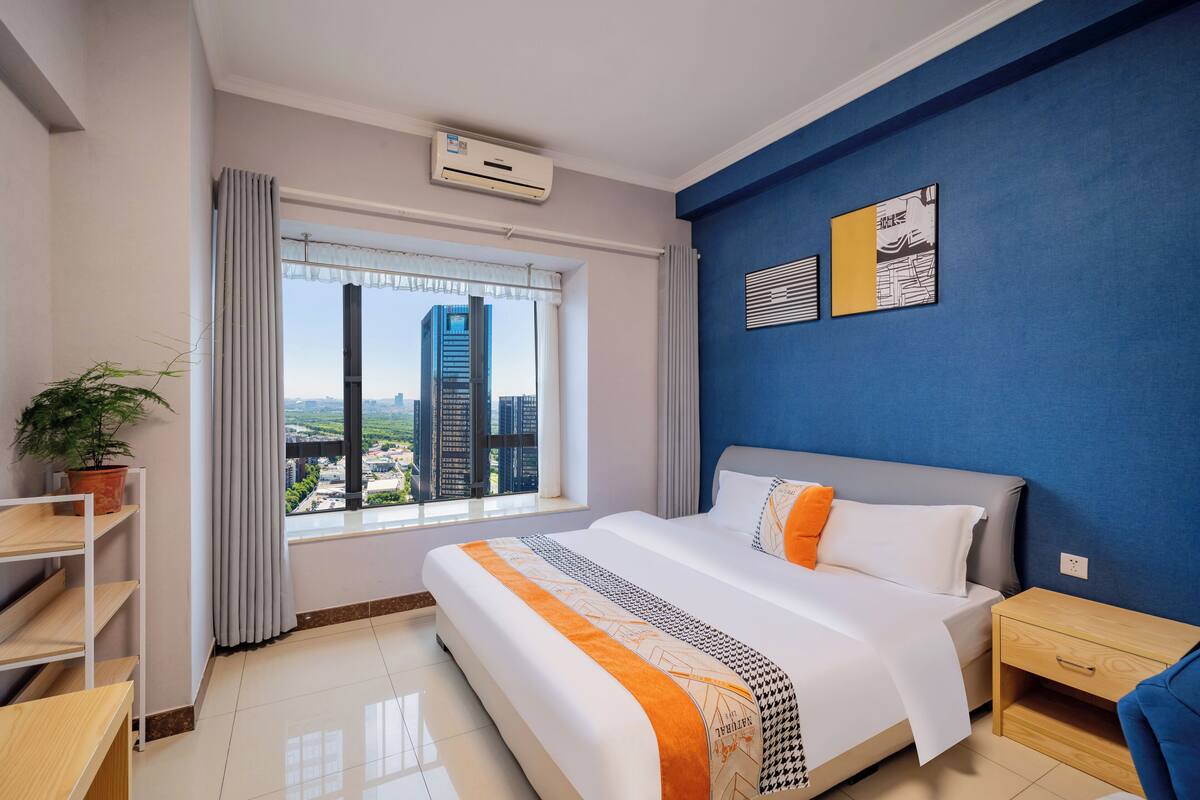 A modern hotel room with a large bed, a view of the cityscape, a potted plant, and a blue accent wall.