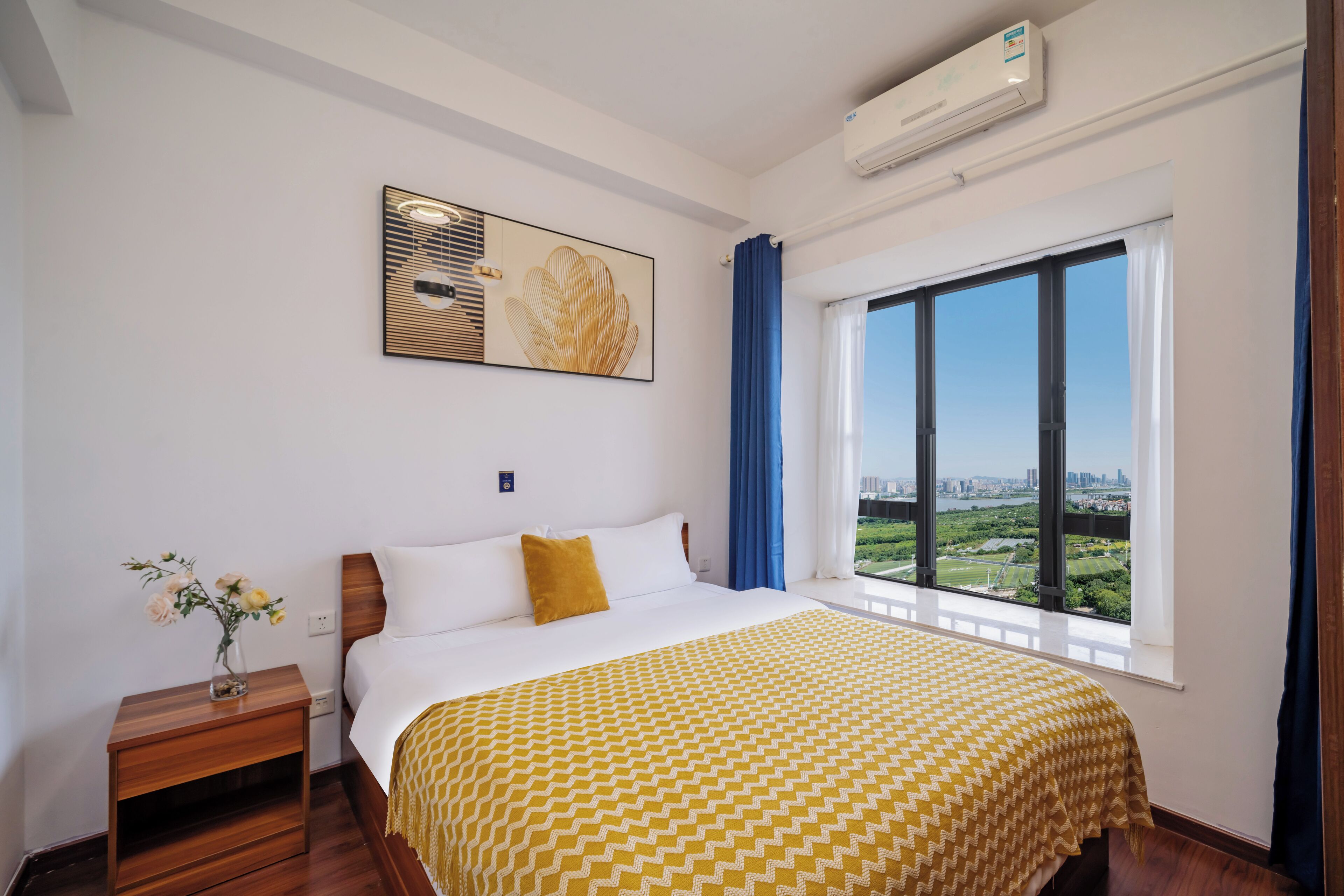 A hotel room with a large window, a bed with a patterned bedspread, a nightstand with a vase of flowers, and a wall-mounted air conditioner.