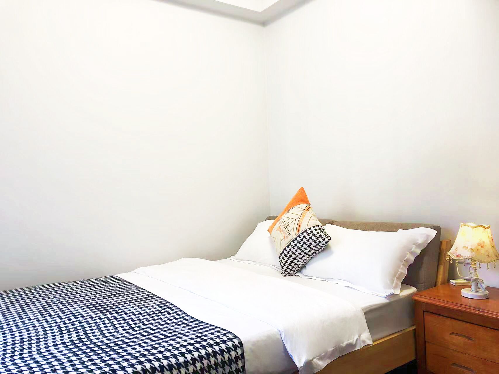 Un lit simple avec une couvre-lit à motifs, deux oreillers, une table de chevet avec une lampe et une petite table de nuit en bois.