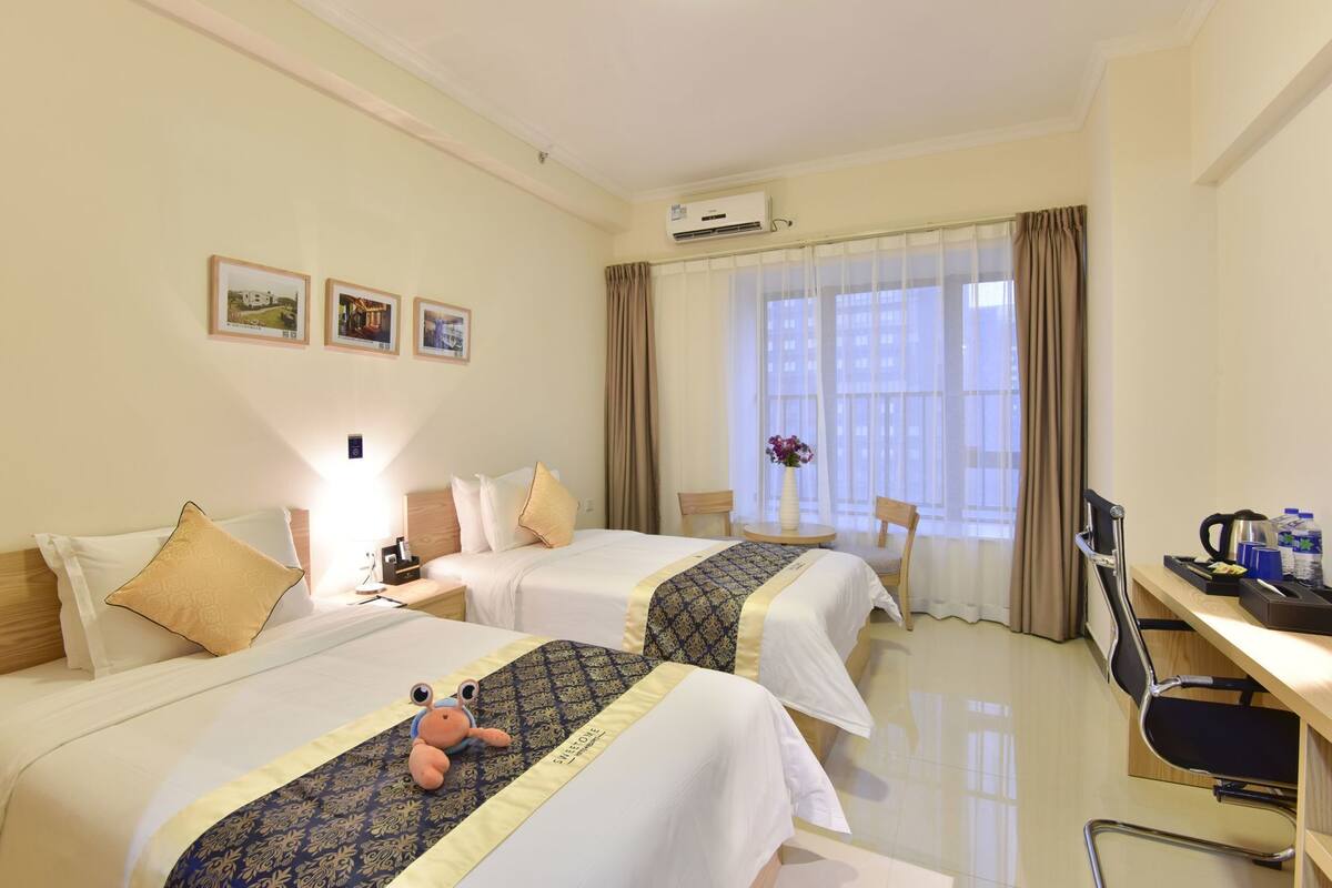 A hotel room with two beds, a desk, a chair, an air conditioning unit, and a window with curtains.