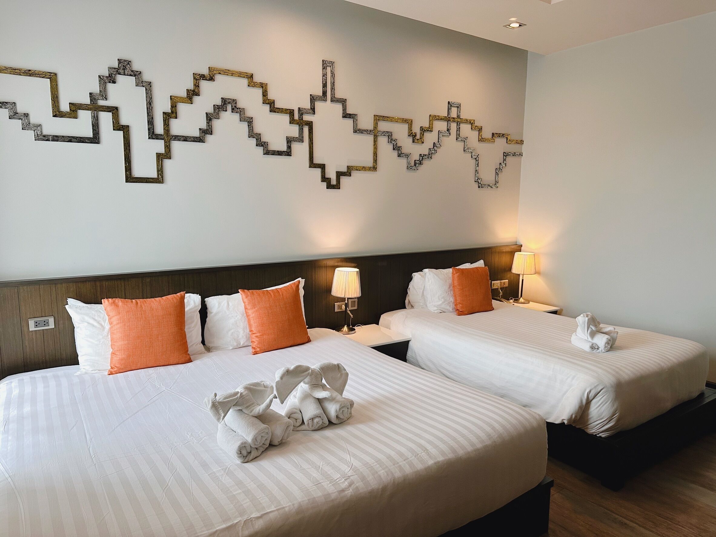 Ein Hotelzimmer mit zwei Betten, jeweils mit weißer Bettwäsche und orangefarbenen Kissen. An beiden Seiten der Betten befinden sich Wandlampen.